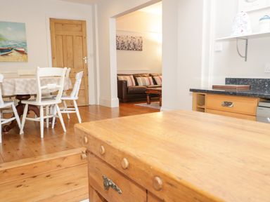 A kitchen area with wooden furniture and white chairs next to a dining table with a checkered tablecloth and a living room with a brown sofa and striped pillows at 3 St Davids Place in Llandudno