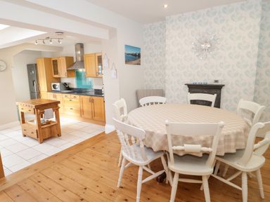 A dining area with a round table and six chairs next to a kitchen with wooden cabinets and an island at 3 St Davids Place in Llandudno