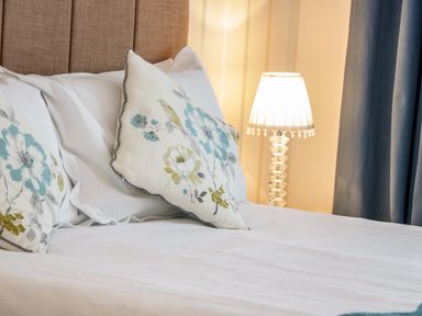 A bed with white linens and floral pillows next to a decorative lamp and blue curtains at 3 St davids Place in Llandudno