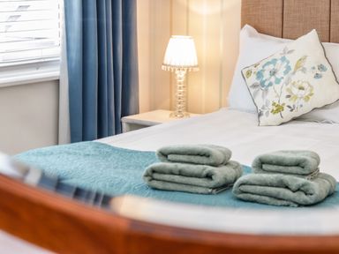 A bed with folded towels on a blanket next to a bedside table with a lamp and a window with curtains at 3 St Davids Place in Llandudno