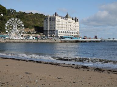A sandy beach with waves near a pier and a large white hotel building with a ferris wheel beside it at 3 St Davids Place in Llandudno