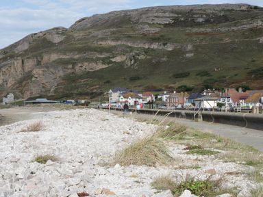 A rocky shoreline with people walking on a paved path and houses at the base of a hillside at 3 St Davids Place in Llandudno