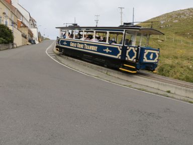 A blue and cream tram car on tracks beside a sloped road with houses on the left and grassy hill on the right at 3 St Davids Place in Llandudno