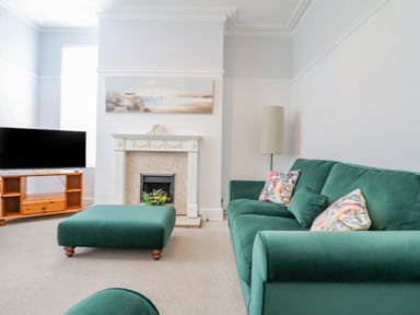 A living room with a green sofa and ottoman a fireplace a wooden TV stand and a floor lamp at 24 Cadnant Park in Conwy