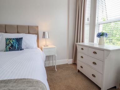 A bedroom with a bed a bedside table with a lamp and a white dresser with flowers by the window at 24 Cadnant Park in Conwy