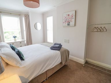 A bedroom with a double bed with pillows and folded towels a dresser with a vase and flowers a round wall mirror and a clothing rack with hangers at 24 Cadnant Park Conwy
