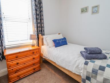 A bedroom with a single wooden bed with white bedding and a blue pillow next to a wooden chest of drawers with a lamp at 24 Cadnant Park in Conwy