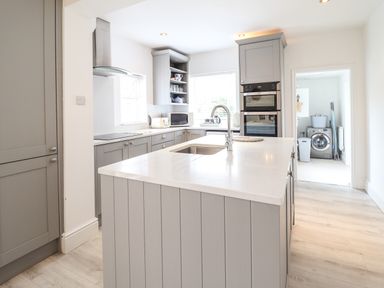 A kitchen with gray cabinets white countertops an island with a sink a double oven and a laundry room visible through a doorway at 24 Cadnant Park in Conwy