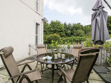 An outdoor patio area with a glass table four chairs a closed umbrella and a garden at 24 Cadnant Park in Conwy