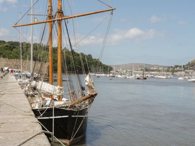 A sailing ship moored alongside a stone dock with many other boats anchored in the water at 24 Cadnant Park in Conwy