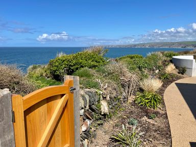 A wooden gate opening to a garden path with shrubs and a view of the sea at The Old Coastguard Lookout in Port Isaac