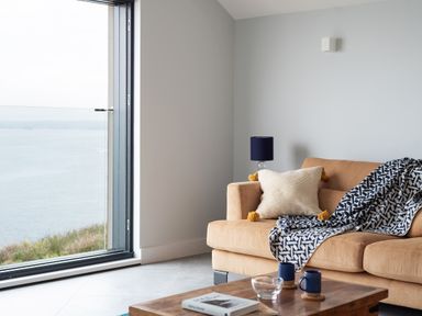 A living room with a tan sofa a pillow a patterned throw a wooden coffee table with mugs and a book and a large window overlooking water at The Old Coastguard Lookout in Port Isaac