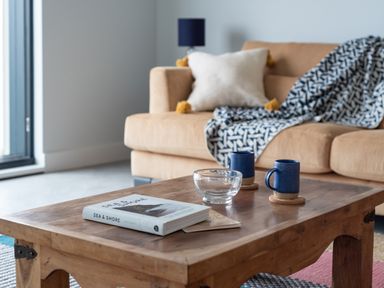 A wooden coffee table with books a glass bowl and two blue mugs in front of a beige sofa with cushions and a patterned throw at The Old Coastguard Lookout in Port Isaac