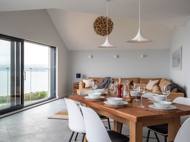 A dining area with a wooden table set with plates, bowls, and glasses next to a living room with a brown sofa and a large glass sliding door at The Old Coastguard Lookout in Port Isaac