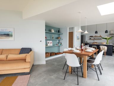 A dining area with a wooden table set for six white chairs and a living room with a tan sofa a light blue built-in shelving unit and a kitchen with black cabinets and hanging lights at The Old Coastguard Lookout in Port Isaac