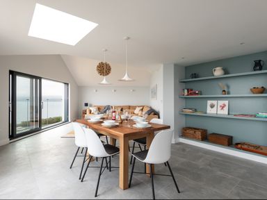 A dining area with a wooden table and white chairs next to a living room with a sofa and open shelves at The Old Coastguard Lookout in Port Isaac