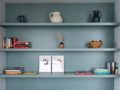 Three teal shelves with books, framed pictures, ceramic jugs, a wooden bowl with pine cones, and a small vase with dried plants at The Old Coastguard Lookout in Port Isaac