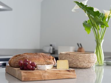 A kitchen counter with bread grapes and two cheeses on a wooden board with a woven basket and flowers in a glass vase at The Old Coastguard Lookout in Port Isaac