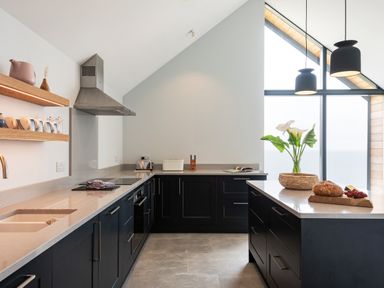 A modern kitchen with dark cabinets white countertops an island with flowers bread and grapes and a large window with a sea view at The Old Coastguard Lookout in Port Isaac