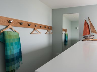 A room with a wooden coat rack with hangers and a green blue checkered scarf a large mirror and a model sailboat on a white surface at The Old Coastguard Lookout in Port Isaac