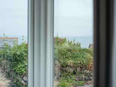 A view through a window showing greenery stone wall rooftops and the sea at The Old Coastguard Lookout in Port Isaac