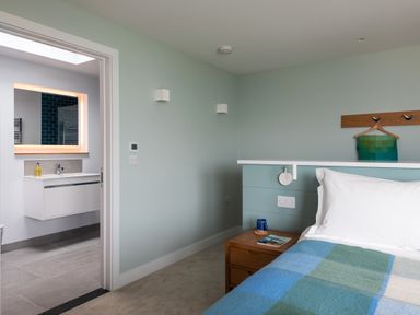A bedroom with a bed featuring white pillow and blue-green checkered blanket a wooden bedside table with a mug and book and an adjacent bathroom with a lit mirror at The Old Coastguard Lookout in Port Isaac