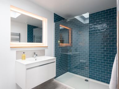 A bathroom with a white sink cabinet and a large mirror with lights above a walk-in shower with blue tiled walls at The Old Coastguard Lookout in Port Isaac