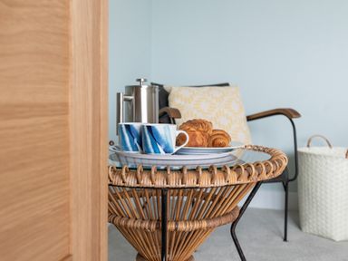 A small table with a coffee pot mugs and croissants next to a chair with a cushion and a laundry basket in a room at The Old Coastguard Lookout in Port Isaac