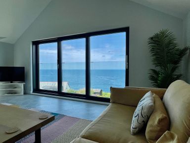 A living room with a beige sofa a coffee table a tv on a white stand and large windows overlooking the sea at The Old Coastguard Lookout in Port Isaac