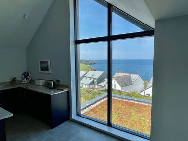 A kitchen corner with a kettle and cutting board next to a large window showing houses and the sea outside at The Old Coastguard Lookout in Port Isaac