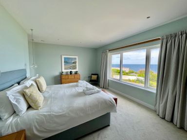 A bedroom with a large bed pillows and folded towels a wooden chest of drawers a chair and a large window with a sea view at The Old Coastguard Lookout in Port Isaac
