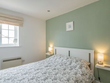 A bedroom with a double bed with floral duvet two bedside lamps a window and a green accent wall at Shrimper's Cottage in Tredrizzick near St Minver