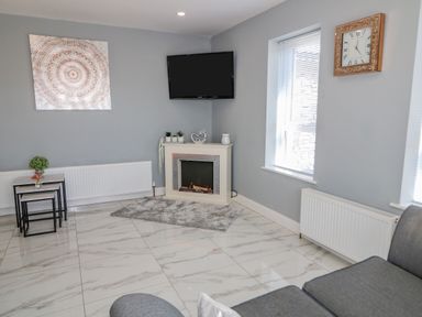 A living room with a gray sofa a wall-mounted television an electric fireplace two windows with blinds nesting tables and decorative plants at SJs Cottage in Castlerock