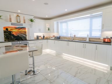 A kitchen with white cabinetry a kitchen island with stools and a large window with blinds at SJs Cottage in Castlerock