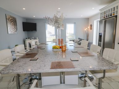 A dining area with a large marble table surrounded by white chairs a television on the wall and a refrigerator in the corner at SJs Cottage in Castlerock