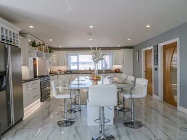 A kitchen with white cabinets a large island with white chairs and marble floor at SJs Cottage in Castlerock