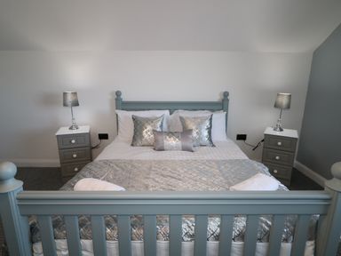 A bedroom with a blue bed frame gray bedside tables with lamps and silver pillows at SJs Cottage in Castlerock