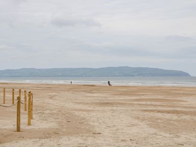A sandy beach with wooden posts and rope fencing leading to the sea with people walking and hills in the background at SJs Cottage in Castlerock