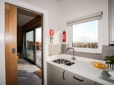 A kitchen sink with a window showing a landscape and a sliding door leading to a wooden room at Hilltop Lodge in Corwen