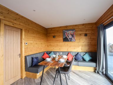 A dining area with a wooden table set with bowls and glasses grey bench seating with colorful cushions wooden walls and a door with glass sliding doors and curtains at Hilltop Lodge in Corwen