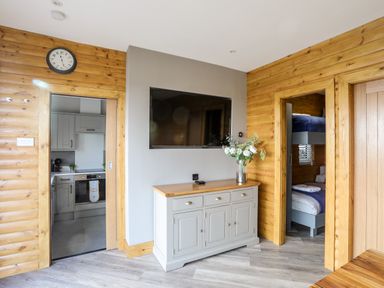 A room with wooden walls a wall-mounted TV a sideboard with flowers a clock and doorways leading to a kitchen and a bedroom at Hilltop Lodge in Corwen