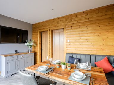 A dining area with wooden table set for four with bowls glasses and plants and a gray sideboard with flowers and a wall mounted TV at Hilltop Lodge in Corwen
