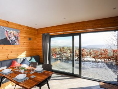 A dining area with wooden table set with bowls glasses and plants next to sliding glass doors showing outdoor patio and landscape at Hilltop Lodge in Corwen