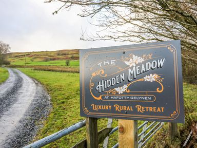 A sign for The Hidden Meadow luxury rural retreat next to a gravel road and green fields at Hilltop Lodge in Corwen