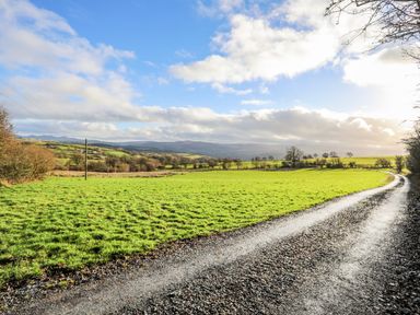 A gravel road next to a green field with trees and hills in the background at Hilltop Lodge in Corwen