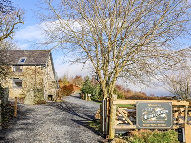 A gravel driveway leading to a stone building with a tree and a wooden gate with a sign for The Hidden Meadow luxury rural retreat at Haptofty Gelynen