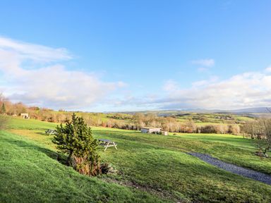 A grassy hillside with picnic tables and a small tree with distant hills and a blue sky at Hilltop Lodge in Corwen