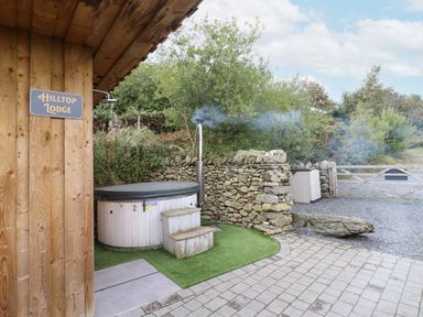 An outdoor area with a hot tub beside a wooden building with a Hilltop Lodge sign and a stone wall in Corwen