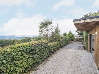 A paved driveway next to a wooden building with trees and hills in the background at Hilltop Lodge in Corwen