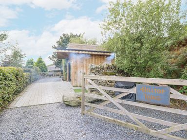 A wooden gate with Hilltop Lodge sign next to a driveway with paving stones and a wooden building surrounded by trees and bushes at Hilltop Lodge in Corwen